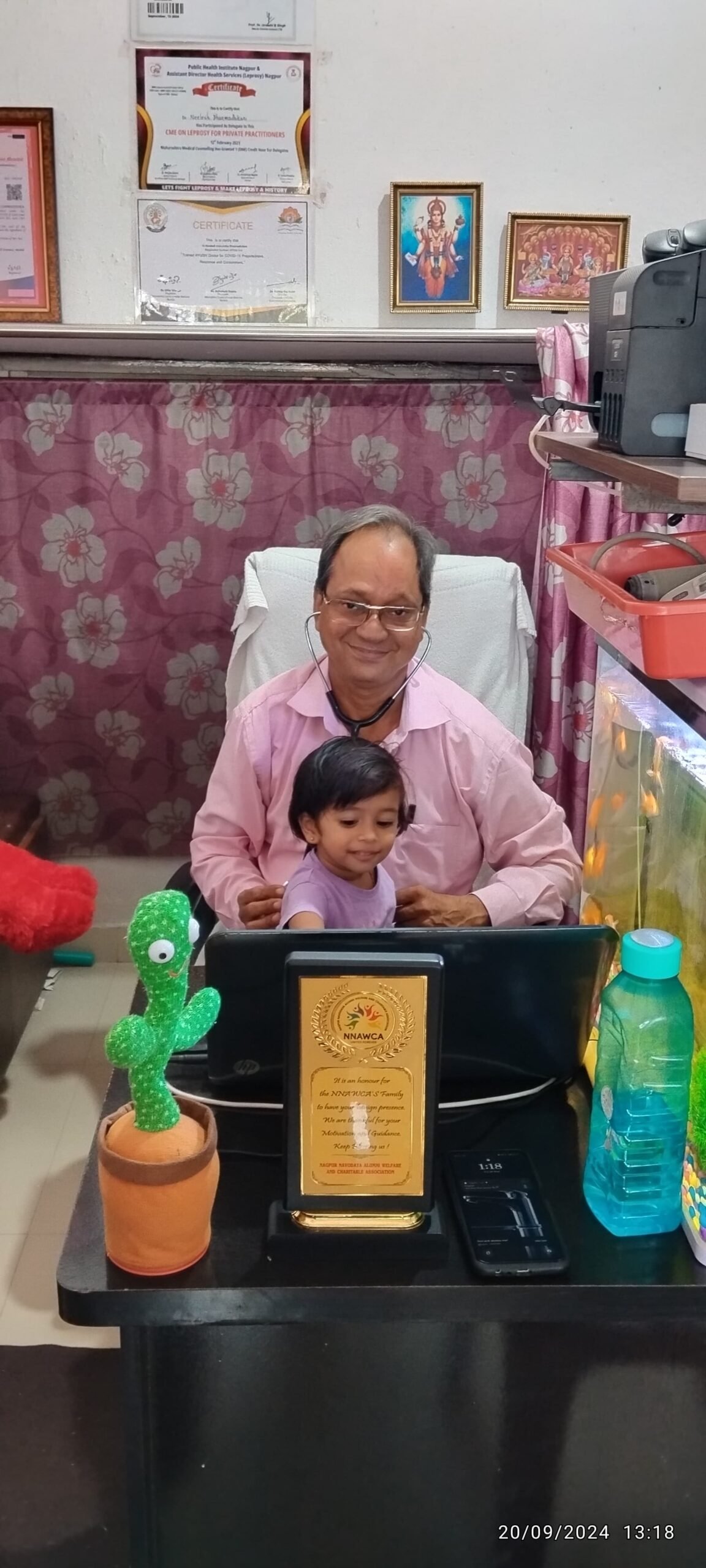 Dr. N. A. Dharmadhikari auscultating the back of a 2.5-year-old baby patient sitting on his lap while she curiously watches the laptop screen