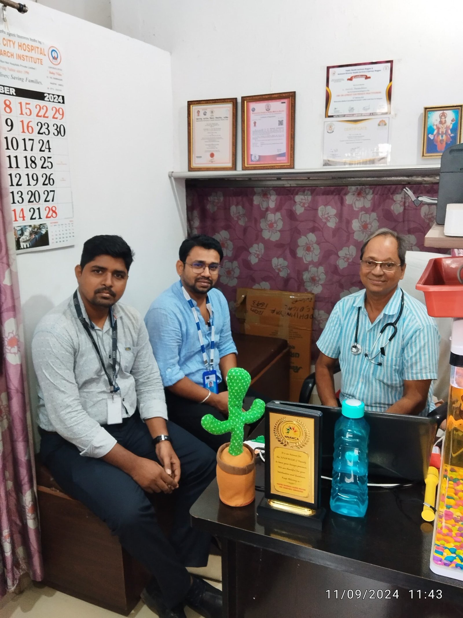 Mr. Biplov Mandal, Mr. Milind Gavhande, and Dr. N. A. Dharmadhikari seated together during a formal visit at the clinic, with certificates on the wall and a plaque on the table.
